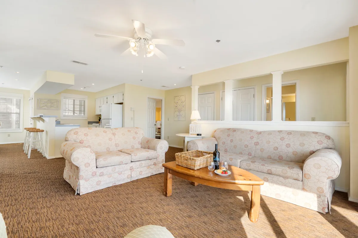 A spacious, cozy living room with comfortable sofas arranged around a coffee table, seamlessly flowing into the kitchen area in the background. The atmosphere is inviting, ideal for gatherings, relaxation, and entertainment.
