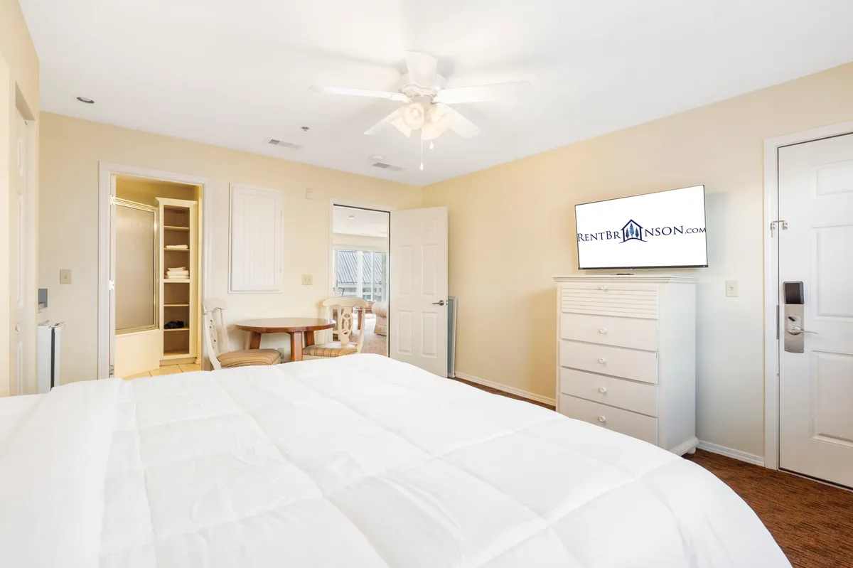A spacious bedroom featuring a comfortable bed, television for in-room entertainment, and a dressing area visible through a nearby doorway. The setup suggests ample space for relaxing, watching movies from bed, and preparing for the day ahead.
