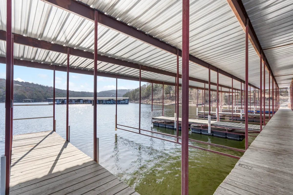 A peaceful lake scene with a dock stretching over the water, boathouses, and a mountainous landscape in the background. The atmosphere is serene, ideal for boating, fishing, or simply relaxing by the lake as the sun sets.