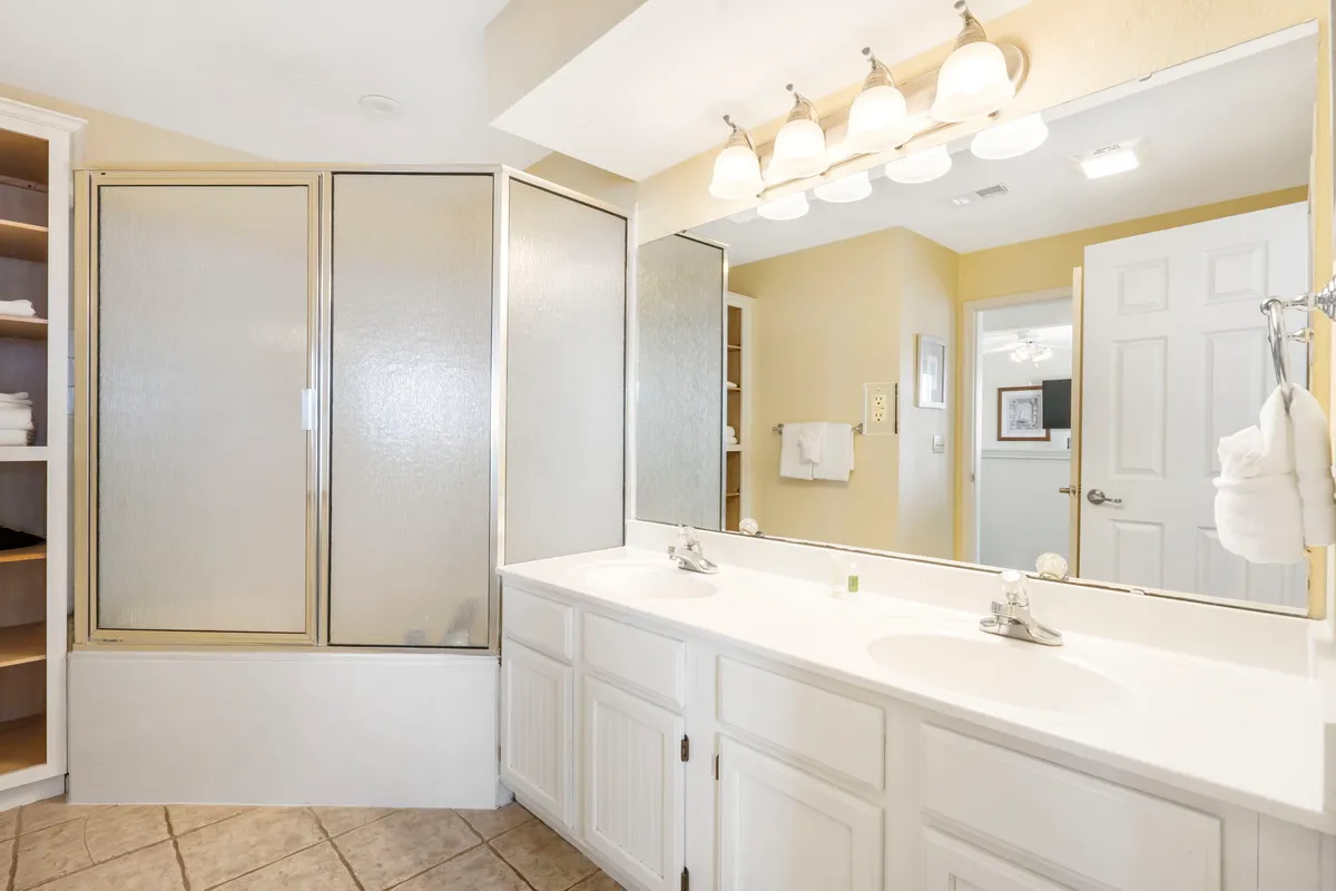 A modern ensuite bathroom featuring a double sink vanity, glass shower enclosure, a large mirror, and open shelving. The space feels bright, refreshing, and organized—ideal for two guests to enjoy their own routines in comfort.