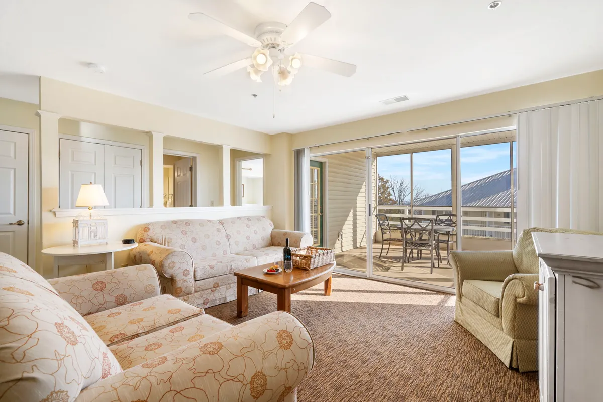 The image showcases a cozy living room with a comfortable sofa and coffee table, leading out to a private patio through a sliding glass door. The patio offers sunset views over Table Rock Lake, setting the scene for relaxation and memorable moments. The warm interior invites guests to unwind, play games, and enjoy quality time together.