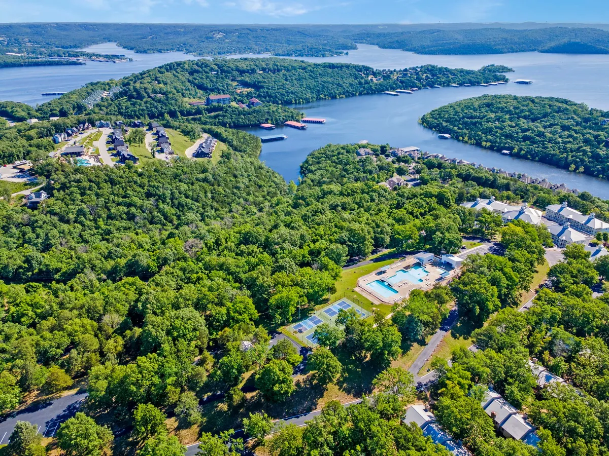 This image presents a panoramic lakeside view of the resort with a swimming pool in the foreground, tennis courts nearby, and dense greenery surrounding well-kept residential buildings. The scene captures the expansive outdoor amenities and their close proximity to the lake.