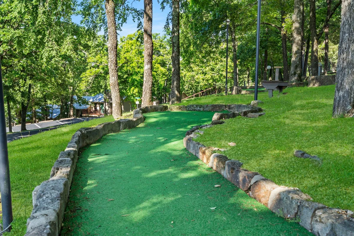 A scenic mini-golf path winds through lush greenery, shaded by tall trees, with benches along the route. The space feels open and inviting, ideal for leisurely walks or playful activities. The area is surrounded by well-maintained lawns and a natural woodland backdrop.