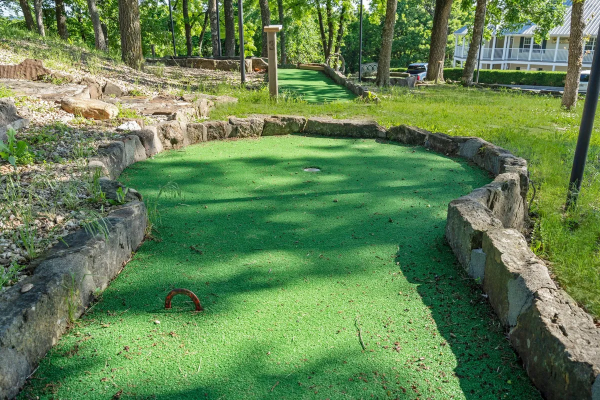 A lively mini-golf course sits surrounded by lush grass and natural stone borders, shaded by trees. Families and friends are depicted enjoying friendly competition in a beautiful outdoor setting.