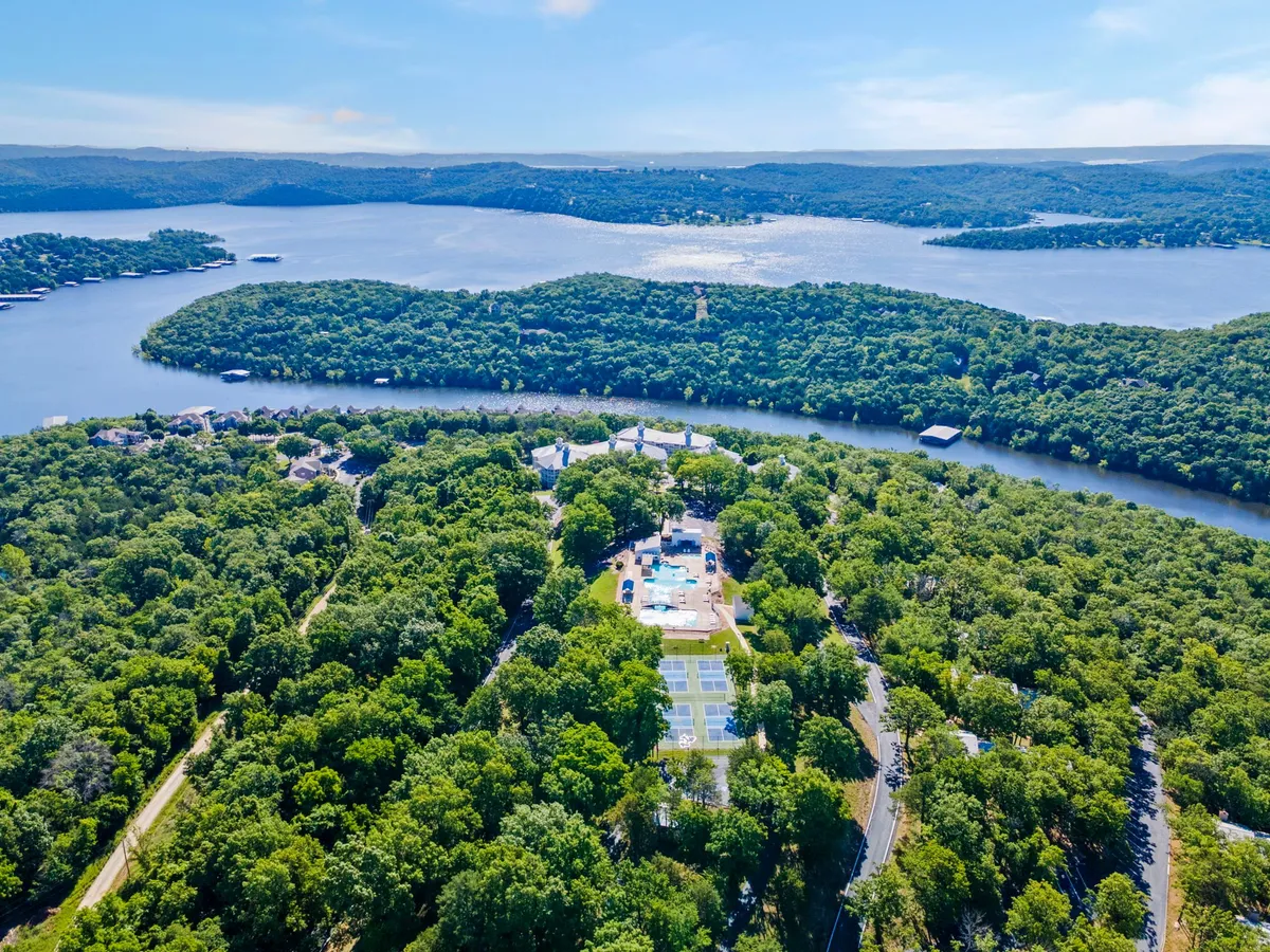 This elevated view captures the resort’s swimming pool, tennis courts, dense greenery, and the expansive lake in the background. The scene conveys a vibrant, inviting atmosphere for outdoor recreation and relaxation, highlighting the proximity of water and land-based amenities.