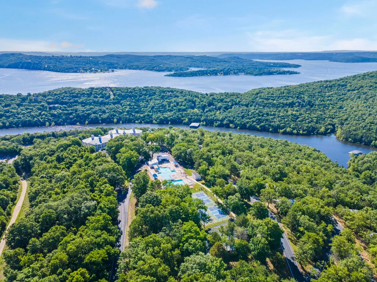 A panoramic view of lush trees, sparkling blue water, a swimming pool, tennis courts, and a winding road weaving through the landscape. The setting is bright and serene, evoking a sense of outdoor freedom and recreational opportunities.