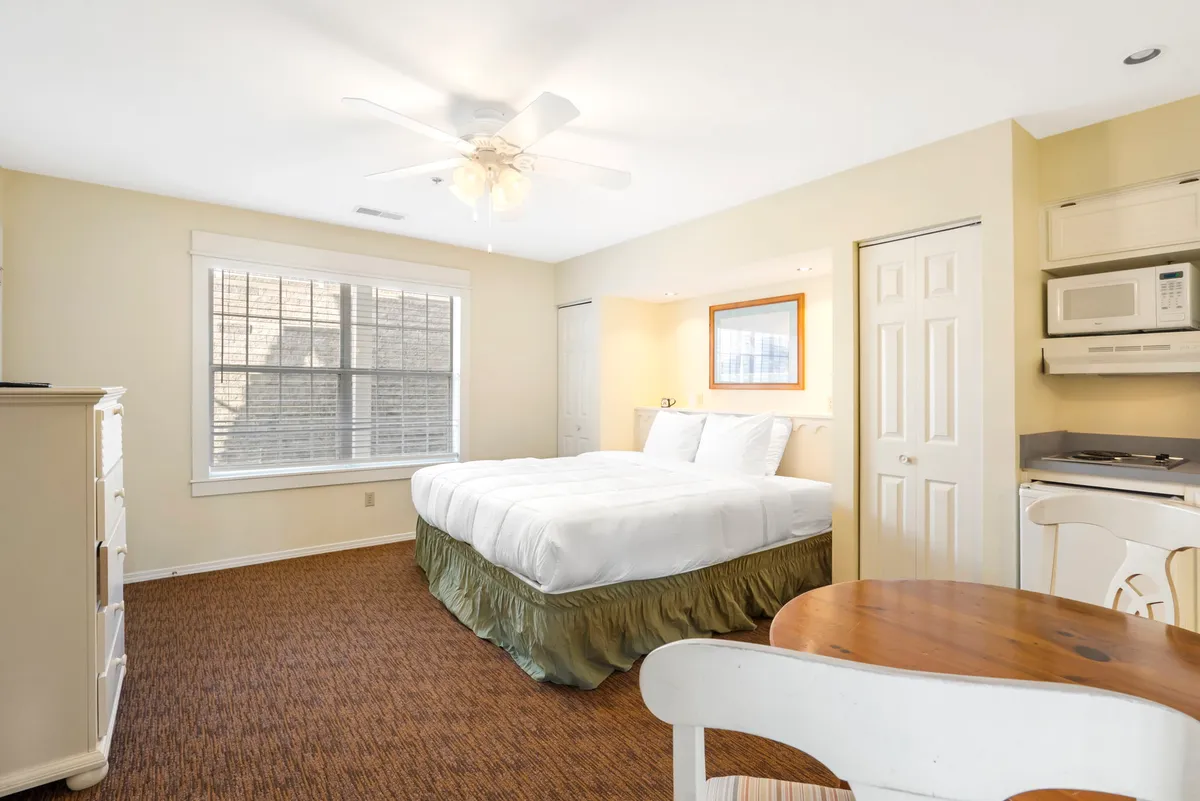 A comfortable bedroom with a queen-sized bed, wooden table, microwave, and a window with blinds that lets in natural sunlight. The setting feels cozy and welcoming, ideal for relaxing with your pet after a day of adventure.
